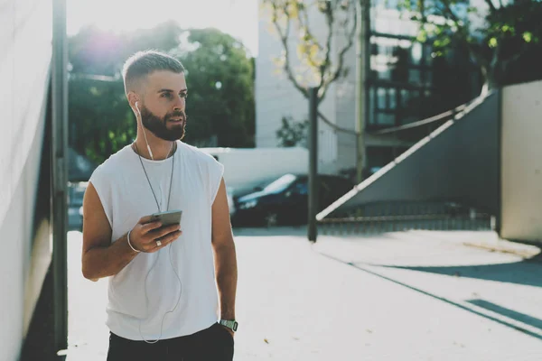 Talented designer reading emails from the clients on a mobile phone while walking the street on a summer sunny day. Hipster bearded guy listening to the music on a smartphone while resting outdoors.