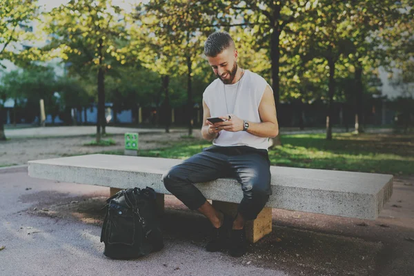 都市公園のベンチに座って携帯電話で旅行ガイド アプリケーションを読む旅行者の笑みを浮かべてください 街を歩きながらスマート フォンで留学生サーフィン Web — ストック写真