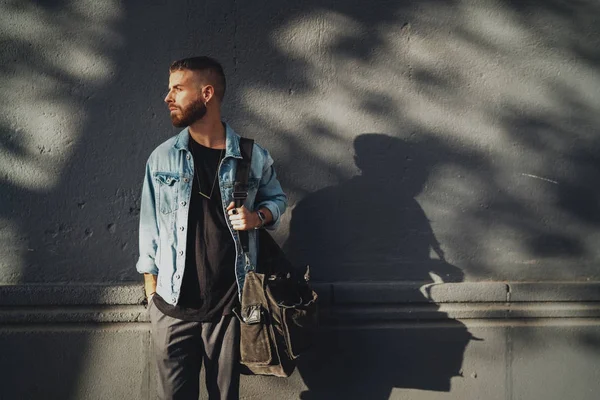 Confident handsome bearded male in jeans jacket posing for the camera on a gray street wall with empty area for logo or design. Stylish young traveler standing outdoors with a bag on a sunny day.