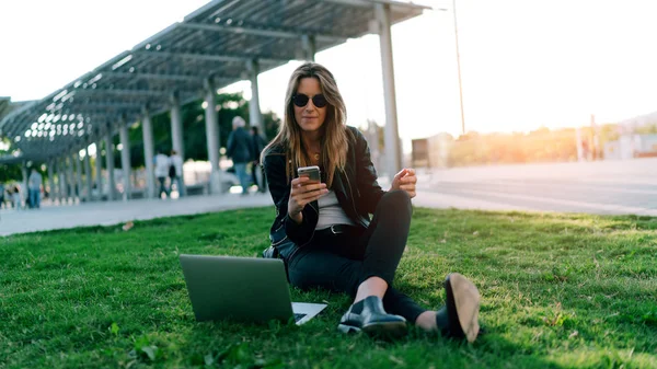 サングラスを身に着けているスタイリッシュな白人女性は ポータブル コンピューターを持って公園に座りながらメールで携帯電話を読んでいます ファッションのブロガーは電子機器によって屋外での作業します — ストック写真