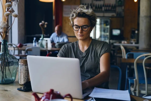 年轻迷人的白种女性戴眼镜是在笔记本电脑上键入信息 连接到 学生女孩正在准备考试 而坐在一个现代化的室内咖啡馆 — 图库照片
