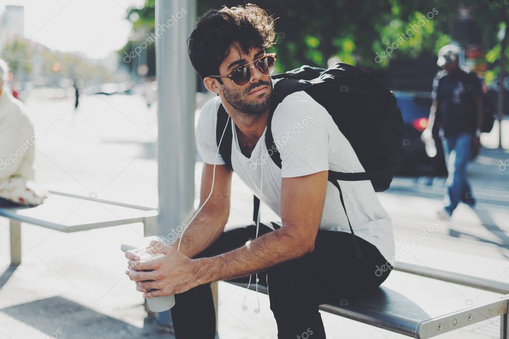 Young bearded man with curly brunette hair wearing sunglasses is waiting for the train on a station. Handsome male is sitting on a bench with a backpack on his back on a blurred urban background.