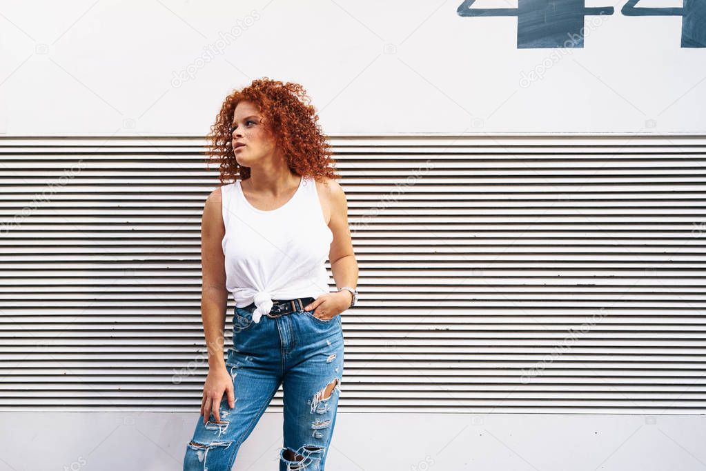 A red-haired hipster girl wearing white t-shirt and jeans is looking aside while standing beside the striped wall. An attractive woman with curly hair and freckle face is standing on the street.