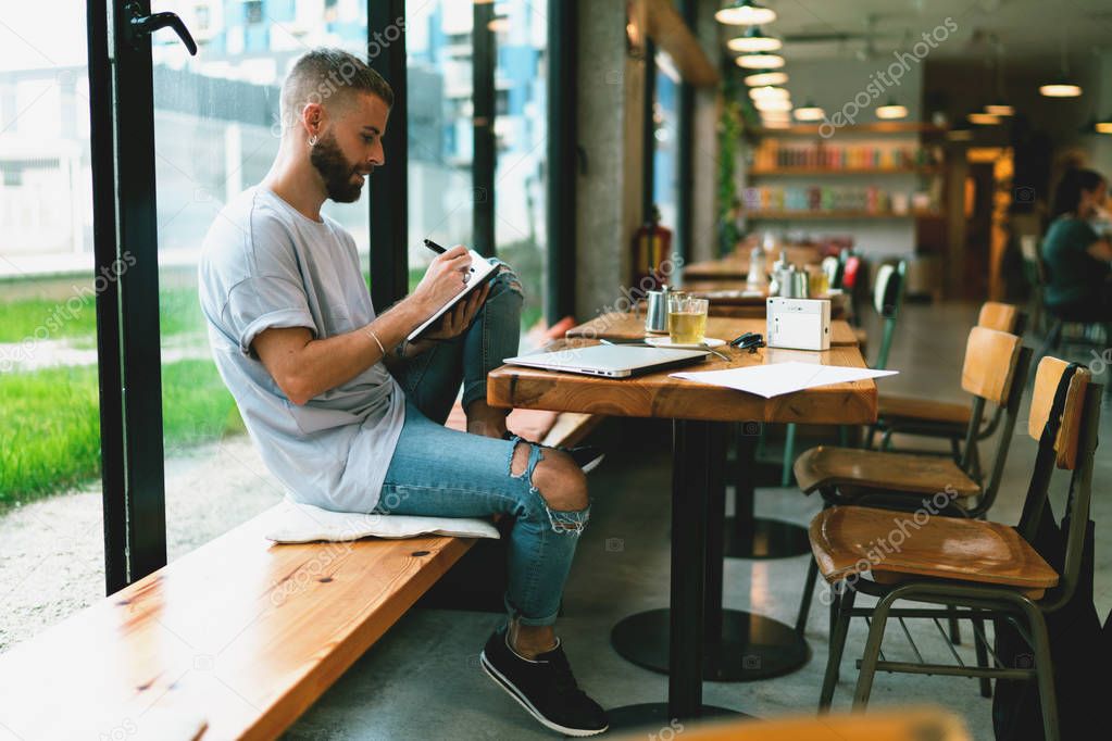 Pensive male blogger making work plan writing main points in notepad while sitting in a modern cafe. Relaxed hipster guy making notes while sitting at the table with a closed laptop on it.