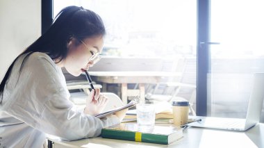 Genç freelancer erkek güneşli bir büyük pencere yanında otururken kitap okuyor. Genç çekici Asyalı kadın yeni sunuya hazırlanırken bilgi arama.