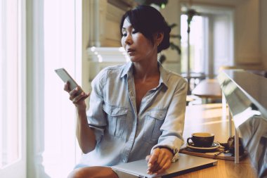 Genç girişimci kadın bir akıllı telefon ve wi-fi için bağlı bir taşınabilir bilgisayar ile bir modern coworking alanda çalışıyor. Asya iş kadın cep telefonu mesajları okuyor.