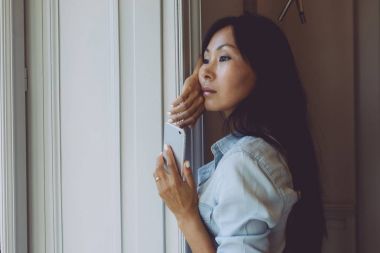 Güzel Asyalı kadın büyük pencerenin önünde bir fincan kahve ile sabah bakarken bir smartphone tutuyor. Güzellik blogger Haberler bir smartphone cep telefonu ile brunch sırasında okuyor.