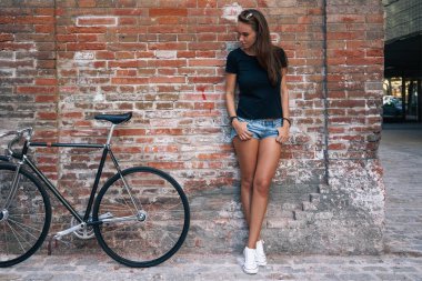 Tuğla duvar arka plan üzerinde duran bir sabit vites bisiklet ile boş siyah t-shirt ve kot şort giyen bir genç sevimli hipster kız açık bir portresi. Metin veya tasarım için boş alan