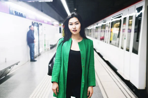 Young beautiful stylish asian lady with long dark hair wearing long green jacket is looking at the camera while standing on a blurred metro platform background while train is coming.