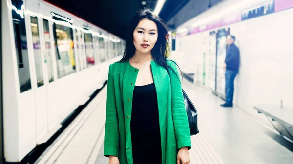 年轻美丽时髦亚洲女性与长的黑头发佩带长的绿色夹克是看在照相机 当站立在一个模糊的地铁站背景 当火车来时 — 图库照片