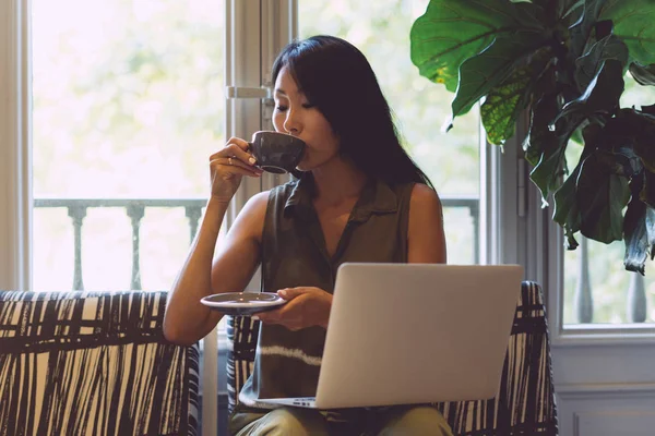 Charming asian woman with long dark hair is drinking coffee while sitting with a portable computer in a modern coffee shop. Young hipster girl is waiting for the friends meetng in a cafe.