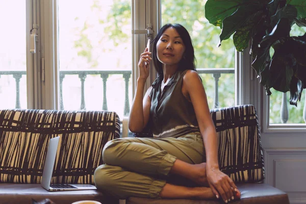 Charming asian hipster girl is talking by smartphone while sitting in a cafe with the modern interior. Attractive business woman wearing casual clothes is making business calls during the coffee break