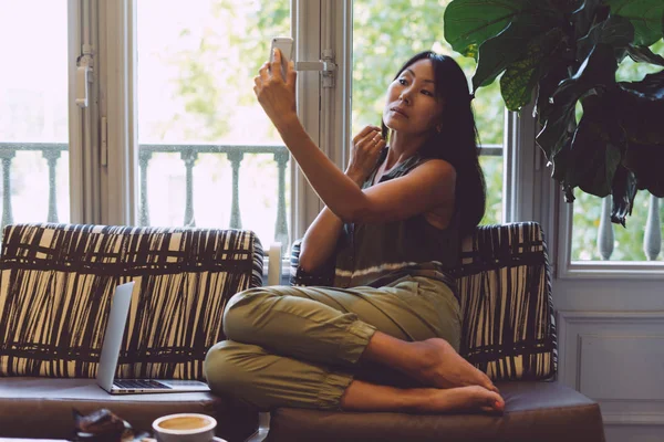 Young beautiful asian woman is making selfie by a mobile phone while sitting on a sofa with a laptop in a modern city cafe. Beauty blogger is looking at the smartphone display to see the new make-up.