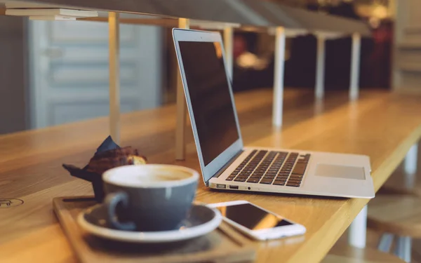 A close up view of a modern portable computer with a blank screen on the blurred cafe background. An opened laptop and mobile phone is placed with a cup of coffee on a table in a restaurant.