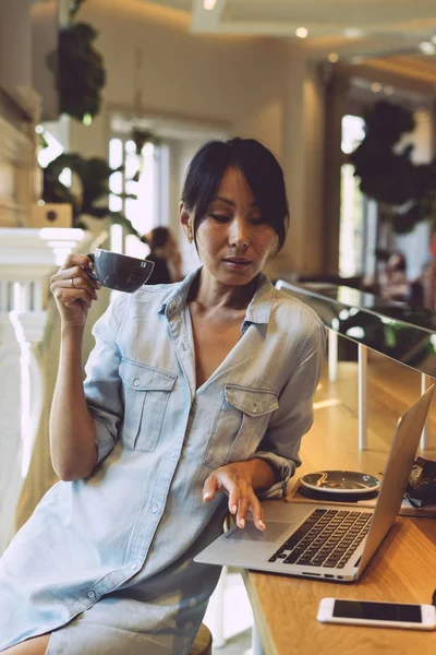 魅力的なアジアの女性は 近代的な都市のカフェで休憩しながら彼女のポータブル コンピューターで Web をサーフィンです 若い美容ブロガーがラップトップで新しいオンライン プロジェクトに取り組んでいます — ストック写真