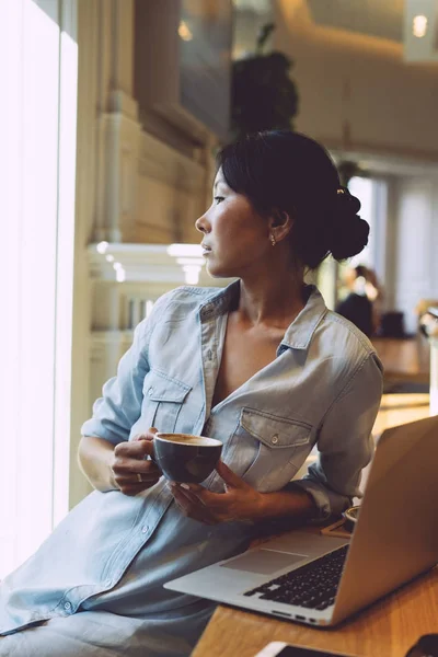 美丽的亚洲女人正看着窗外 坐在桌子上 手持一台手提电脑 年轻的网页设计师是站在一杯茶 而思考一个新的项目 — 图库照片