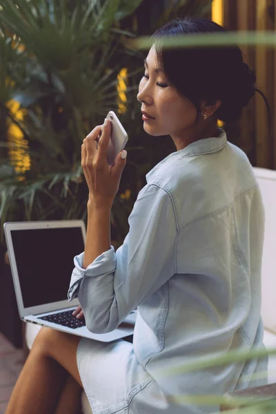 Young woman with dark hair wearing jean dress is looking at the display of her smartphone while opened modern portable computer with blank monitor is lying on her knees. Mock up. Empty space for logo