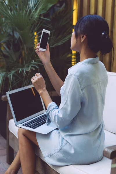 黑头发的年轻女子穿牛仔礼服是看着她的智能手机的空白显示 而打开现代便携式电脑与空的空间是躺在她的膝盖 — 图库照片