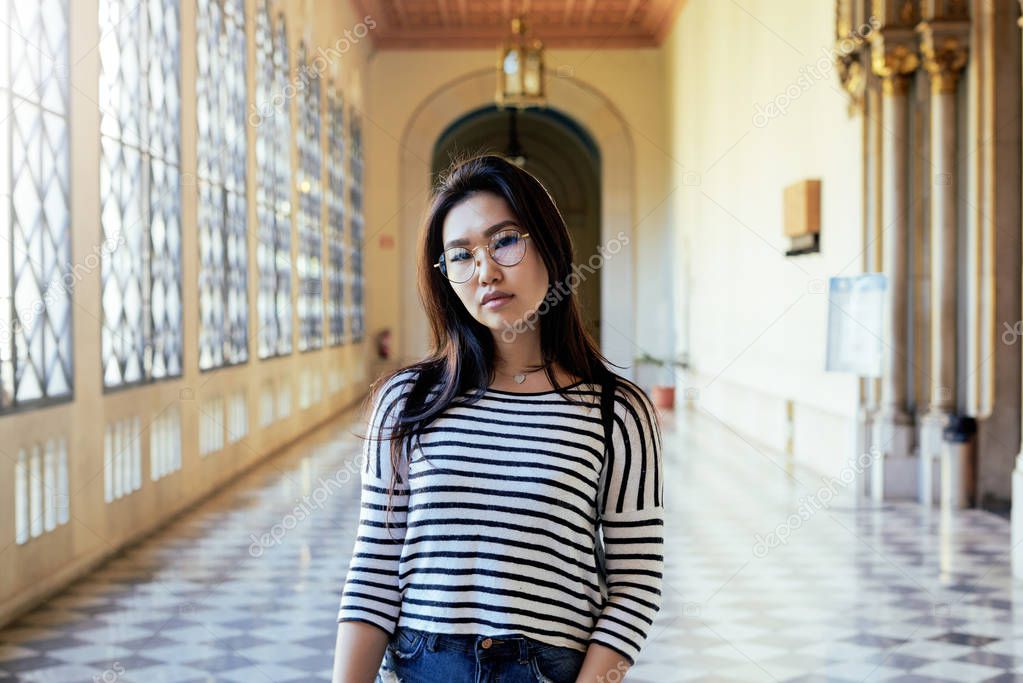 Asian student girl wearing glasses is looking at the camera while standing on the vintage hall background. A hipster female wearing casual clothes is walking the old city historic building.
