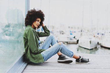 Genç çekici melez kadın kot tulum ve yeşil gömlek giyen afro saç modeli ile bir kenara bir günbatımı üzerinde bulunan şehir yatak alan otururken gülümsüyor. Mutlu hippi kız rahat kıyafetler içinde.