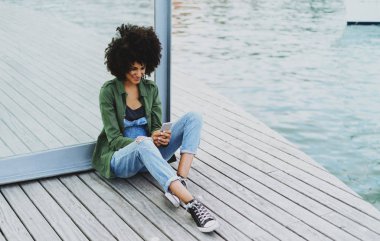 Çekici öğrenci Bayan kot tulum giyen afro saç modeli ile gülümseyen iletileri bir günde şehir rıhtım bölgesinde bir bankta otururken bir ekranda bir cep telefonunun yazmakta olduğunu.