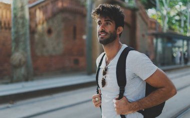 Backpacker iyi sıcak güneşli bir günde şehir merkezine yürüyüş boş zamanlarını mutlu gülümseyerek. Şehir merkezinde bir toplu taşıma için bir yaz günü bekleyen bir sırt çantası ile sakallı Gezgin.