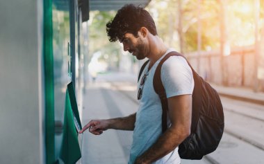Sakallı yakışıklı hipster adam sırt çantası ile rahat kıyafetler giymiş Avrupa şehri tatil gezisi sırasında self servis satış makinesi istasyonu, otobüs biletleri satın alıyor. Işık parlaması.