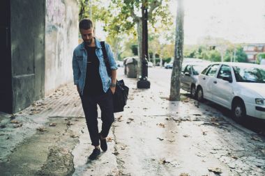 Düşünceli yakışıklı erkek kot ceketli şehir street güneşli bir sonbahar gününde yürüyüş sakallı. Şık genç uluslararası öğrenci açık havada omzunda bir sweekend günde bir çanta ile gezinme.