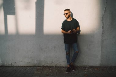 Beyaz duvar arka plan bir sokakta üzerinde duran bir akıllı telefon tutarak sakallı yakışıklı olan. Şık adam waitinig şehri için arkadaş için süre web sörf güneş gözlüğü takıyor.