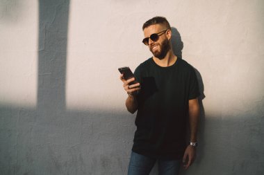 Bir cep telefonu üzerinde girişimci adam okuma e-postalar sokakta yürürken gülümseyerek. Şık hipster beyaz beton duvar arka plan üzerinde duran bir akıllı telefon mesajları yazarak.