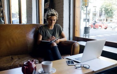 Güzel sarışın rahat kıyafetler giymiş kısa sarı saçlı kadın o üstünde a laptop ile modern bir kafede masada bir vintage deri kanepede otururken bir not defterinde yazıyor.