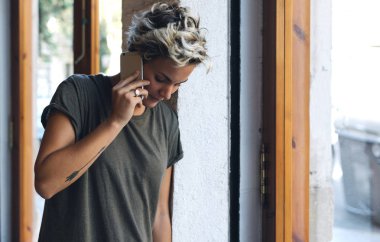 Sarı saçlı genç güzel beyaz kadın yanında bir smartphone pencerenin yanında dururken konuşuyor. Bir haki hippi kız arkadaşlarıyla bir cep telefonu tarafından t-shirt sohbet.