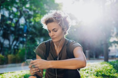 Rahat kıyafetler giymiş sarışın bir beyaz kadın doğal bir arka plan üzerinde otururken bir akıllı telefon mesajları okuyor. Serbest meslek sahibi şehir Garden'da dinlenirken müşteriye mesaj atıyor.