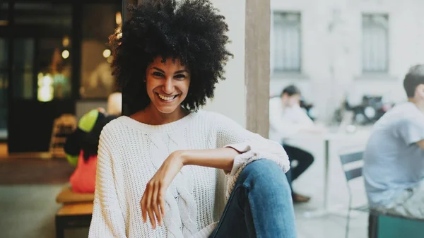 Half length portrait of a cheerful mulatto female with afro hair style wearing stylish clothes smiling at the camera while sitting in a modern city coffee shop on a weekend day on a blurred backgrond.