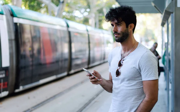 International student with a mobile phone waiting for the bus standing on a blurred urban background. Pensive blogger using public wi-fi on smartphone to find public transport timetable.