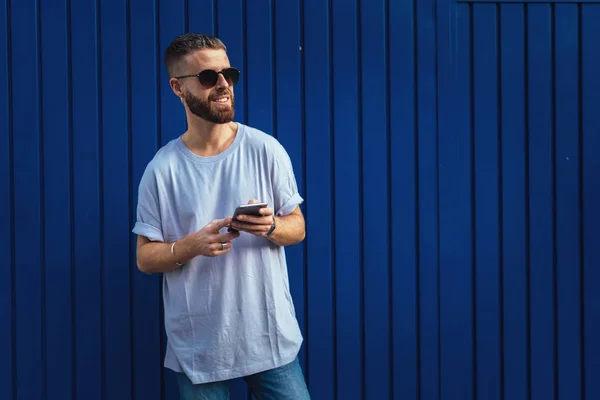 屋外を散歩しながら 携帯電話で友人にスタイリッシュな流行に敏感な男のテキスト メッセージの半身像 青の通り壁に立っている間 Smarthone Web サーフィン髭の男 — ストック写真