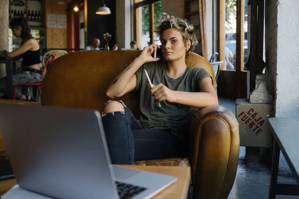若い起業家の女性は コーヒー ショップに座って携帯電話で話してる 金髪の内気な少女はスマート フォン ポータブル コンピューターで テーブルに座って呼び出し元します — ストック写真
