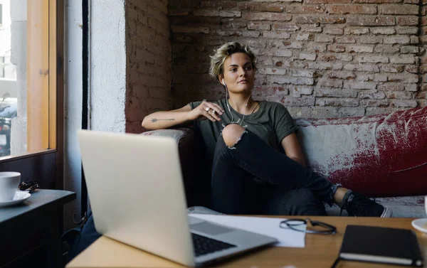 若い女性は 大きな窓の横にあるビンテージ革ソファーのカフェに座りながら新しいプロジェクトのアイデアを考えています ブロンドの女の子は に接続されたラップトップでオンラインで勉強します — ストック写真