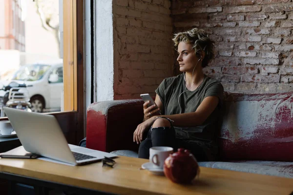 魅力的な女性は ヴィンテージの革のソファに座って携帯電話の画面でメールを読んでいます 若いブロガーがモダンなコーヒー ショップで朝食をとりながら新しい記事を書く — ストック写真