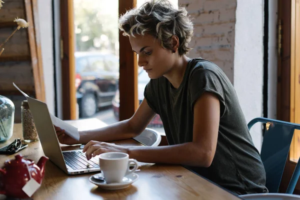 Hipster girl is typing emails by a modern portable computer while sitting in a coffee shop. Young entrepreneur female is waiting for the skype call while having breakfast in a modern cafe.