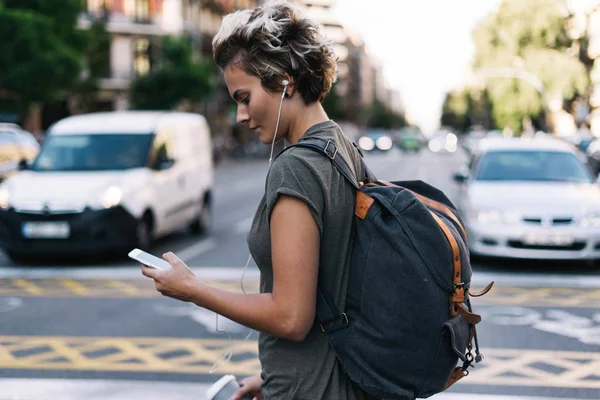 Young blonde woman is walking the city streets with the travel apps on her mobile phone. Hipster girl is typing messages by a smartphone while standing on blurred city street background.