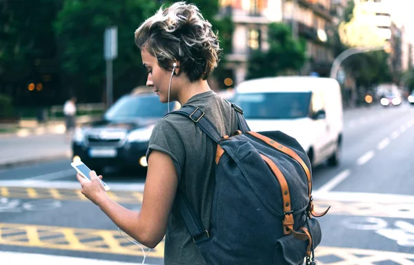 Young hipster girl is exploring new city sights with the travel apps on her mobile phone. Student girl is chatting online by a smartphone while standing on blurred city street background.