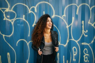 Uzun kıvırcık saçlı mavi duvar grafiti ile bir arka plan üzerinde poz bir siyah deri ceket çekici gülen kızı