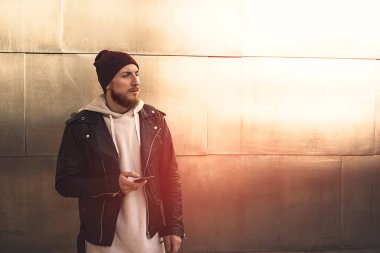 Bir metal duvar arka plan üzerinde beyaz boş sweatshirt ve smartphone ile deri ceket ayakta giyen genç adam