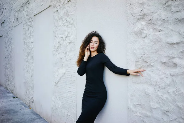 白い壁の背景に電話で話している黒のドレスを着て長い巻き毛を持つ若い笑顔の美しい女性 — ストック写真