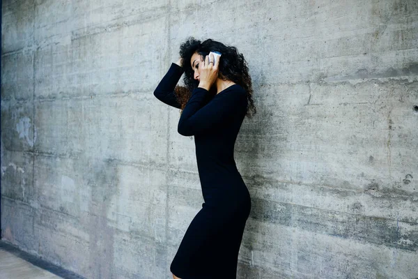 黒のドレスを着て 白いレンガ壁背景に電話で話している長い巻き毛を持つ若い美しい実業家 — ストック写真