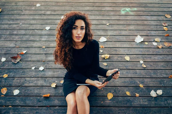 木製の床の背景に公園でリラックスしながらデジタル タブレットを使用して長い巻き毛を持つ若い魅力的な女性 — ストック写真