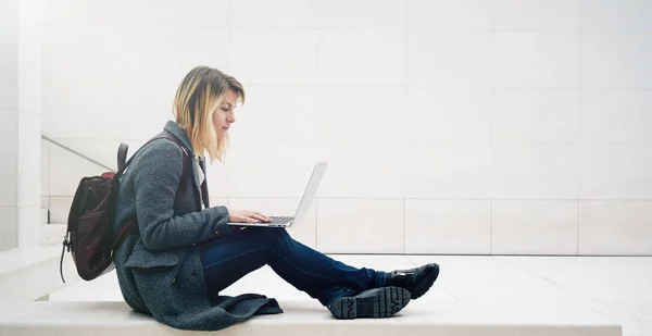 young attractive blonde female student learning online on her laptop while sitting on the stone bench