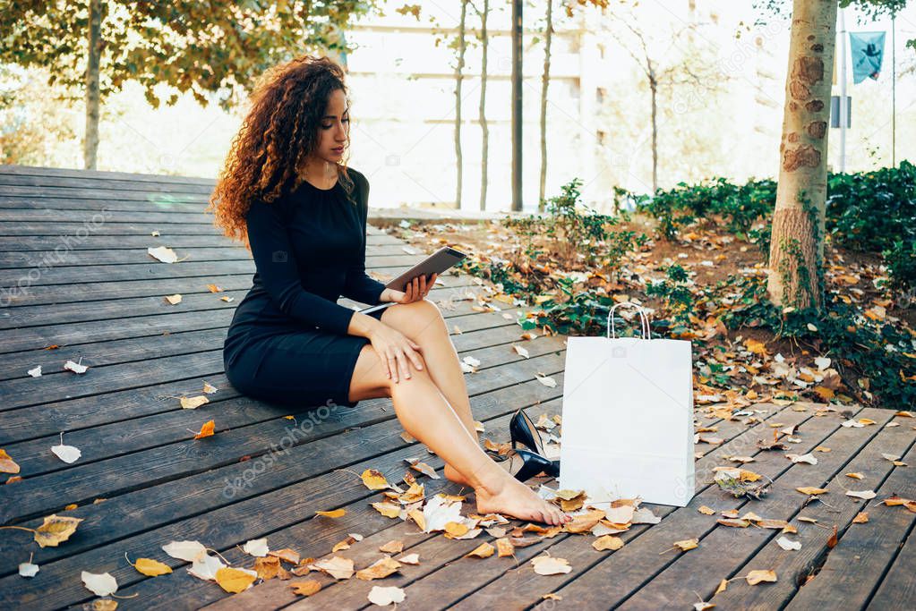 young beautiful woman with long curly hair making online purchases during the lunch break while sitting in the park with white blank paper bag standing next to her