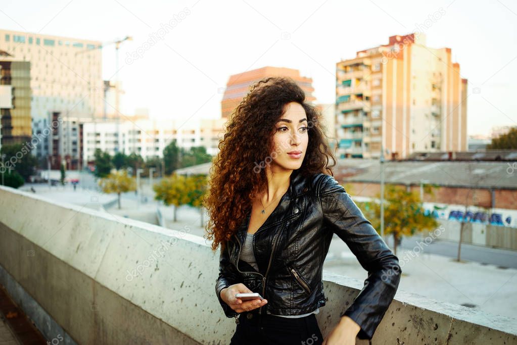 portrait of a cute brunette woman with long curly hair wearing a black leather jacket holding smartphone on a city buildings background
